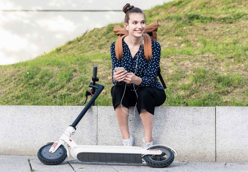 Junge Frau sitzt auf einer niedrigen Mauer. Vor ihr liegt ein E-Scooter.