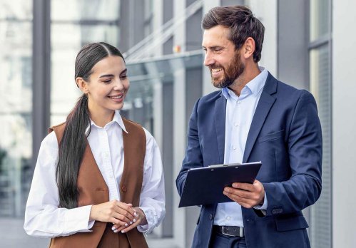 Mann und Frau in Business-Kleidung schauen außerhalb eines Bürogebäudes auf ein Tablet.