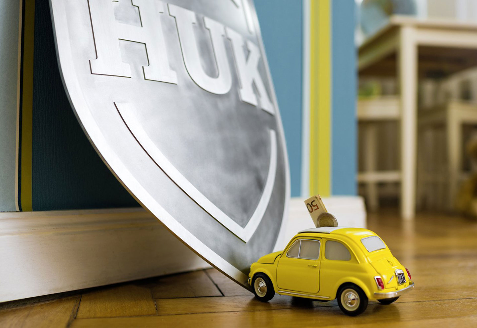 Ein kleines gelbes Auto steht vor dem HUK-Schild. Das Auto ist eine Spardose.