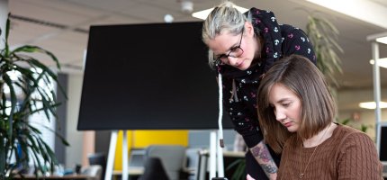 Eine Person steht und eine andere sitzt in einem Büro, umgeben von Pflanzen. Im Hintergrund ist eine leere Tafel zu sehen.