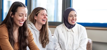 Drei Personen sitzen in einem hellen Raum mit großen Fenstern und unterhalten sich.