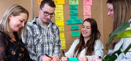 Personen arbeiten im Büro vor einer Wand mit bunten Notizzetteln. Sie halten Stifte und Papier. Eine Pflanze ist im Vordergrund.