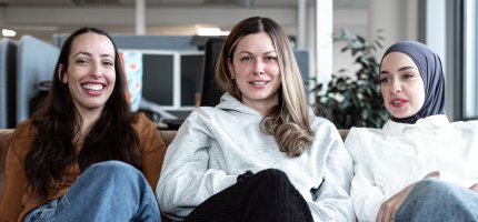 Drei Personen sitzen entspannt auf einem Sofa in einem Büro. Im Hintergrund sind Arbeitsplätze und Pflanzen zu sehe