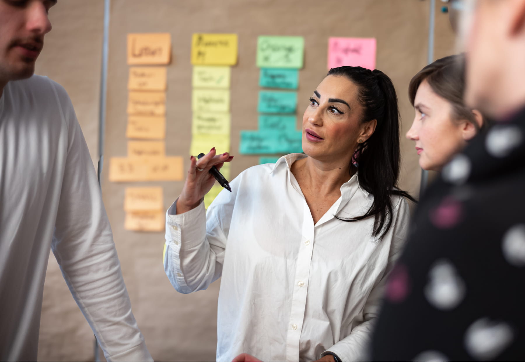 Personen diskutieren vor einer Wand mit bunten Notizzetteln. Eine Person hält einen Stift in der Hand.