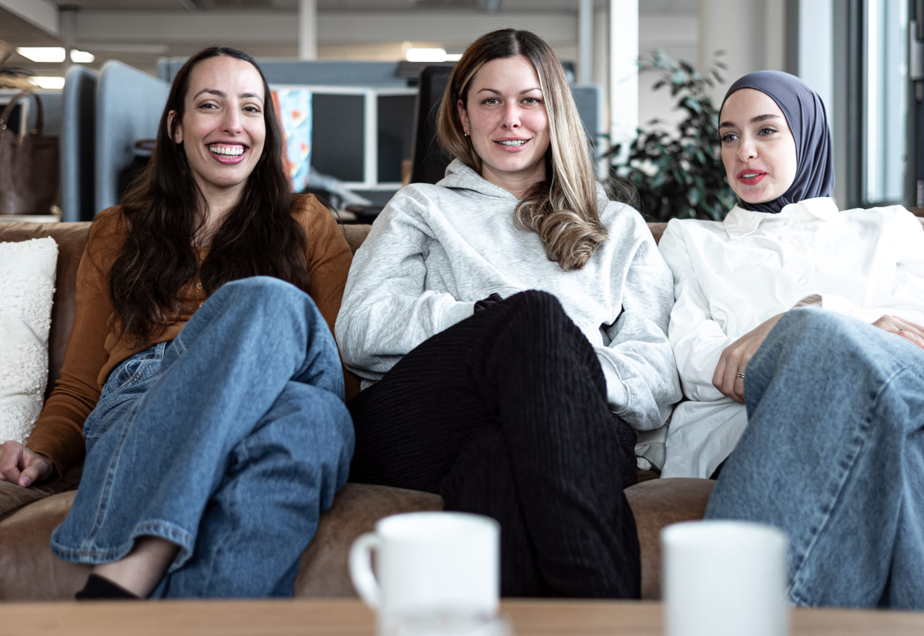 Drei Personen sitzen auf einem Sofa im Büro. Im Vordergrund stehen Tassen.