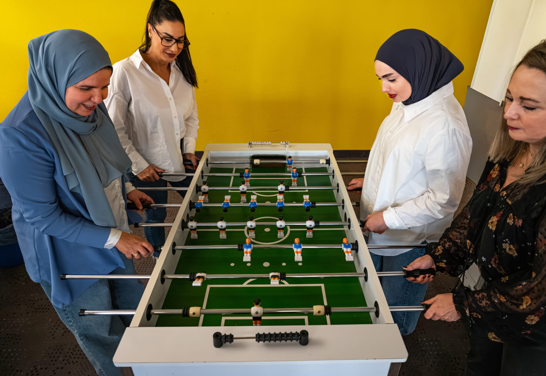 Vier Personen spielen Tischfußball vor einer gelben Wand.