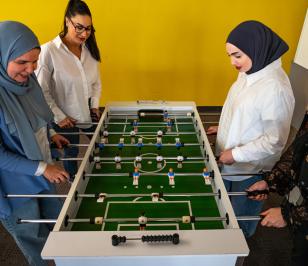 Vier Personen spielen Tischfußball vor einer gelben Wand.