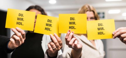 Vier gelbe Zettel mit der Aufschrift: DU. WIR. HDL. werden von Mitarbeiterinnen in die Luft gehalten.
