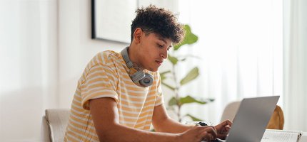 Ein junger Mann sitzt mit Kopfhörern an einem Tisch und arbeitet am Laptop, umgeben von Pflanzen und hellem Licht.