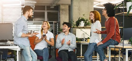 Eine Gruppe von fünf Personen sitzt und steht in einem Büro, unterhält sich und gestikuliert lebhaft. Der Raum ist modern eingerichtet mit Computern und Pflanzen.