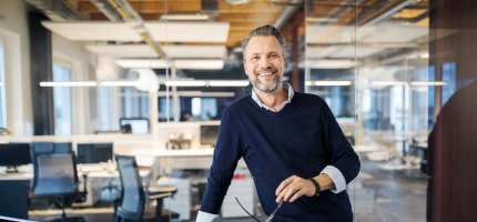 Ein Mann im Büro steht lässig mit einem dunkelblauen Pullover und hält eine Brille. Im Hintergrund sind moderne Arbeitsplätze und große Fenster sichtbar.