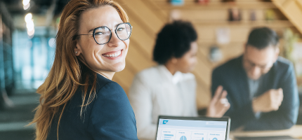 Eine Frau sitzt am Laptop und lächelt in die Kamera. Im Hintergrund sind zwei Personen zu sehen, die sich unterhalten.