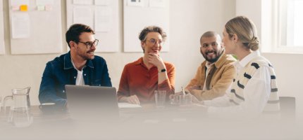 Eine Gruppe aus vier Personen sitzt in einem hellen Besprechungsraum an einem Tisch. Vor ihnen stehen ein Laptop, Notizbuch und mehrere Wassergläser sowie eine Karaffe. Im Hintergrund hängen Whiteboards, und von rechts fällt Tageslicht durch ein großes Fenster. Die Personen wirken konzentriert und im Gespräch vertieft.