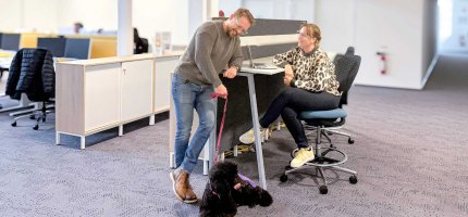 Eine Frau sitzt an einem Schreibtisch und spricht mit einem Mann, der daneben steht und eine schwarze Pudelhündin an der Leine hält. Beide tragen lässige Kleidung; auf dem Tisch steht ein Laptop.
