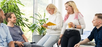 Menschen versammeln sich in einem hellen Raum, einige halten Notizen und Getränke in der Hand.