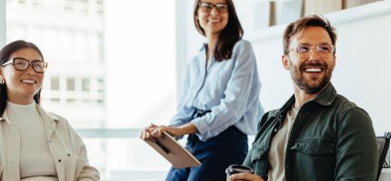 Drei Personen befinden sich in einem Büro, eine sitzt mit einer Kaffeetasse, eine andere steht mit einem Klemmbrett. Der Raum ist hell und modern eingerichtet.