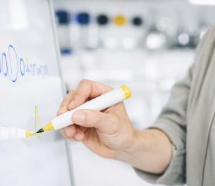 Eine Person schreibt mit einem gelben Marker auf eine weiße Tafel.