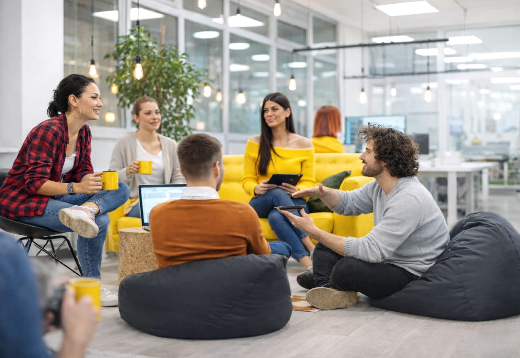 Personen sitzen in einem modernen Büro, trinken aus Tassen und arbeiten mit Laptops und Tablets. Im Hintergrund sind Pflanzen und helle Beleuchtung.