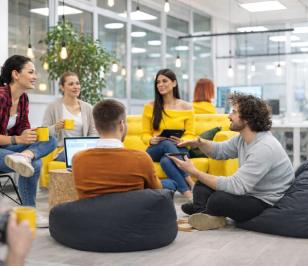 Personen sitzen in einem modernen Büro, trinken aus Tassen und arbeiten mit Laptops und Tablets. Im Hintergrund sind Pflanzen und helle Beleuchtung.