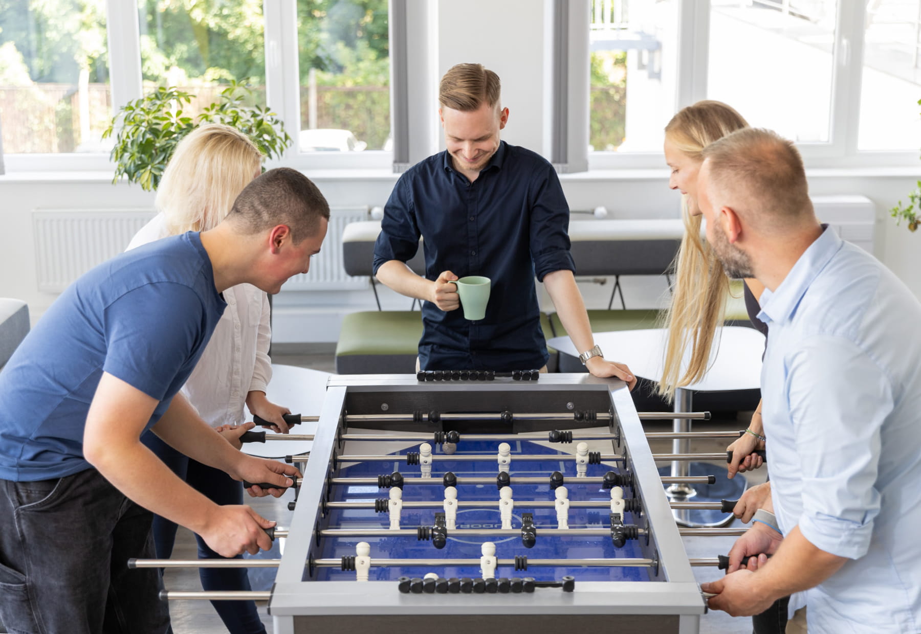 Personen spielen Tischfußball in einem hellen Raum. Eine Person hält eine Tasse.