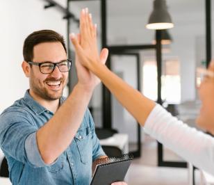 Zwei Personen geben sich ein High-Five in einem Büro. Eine Person hält ein Notizbuch.