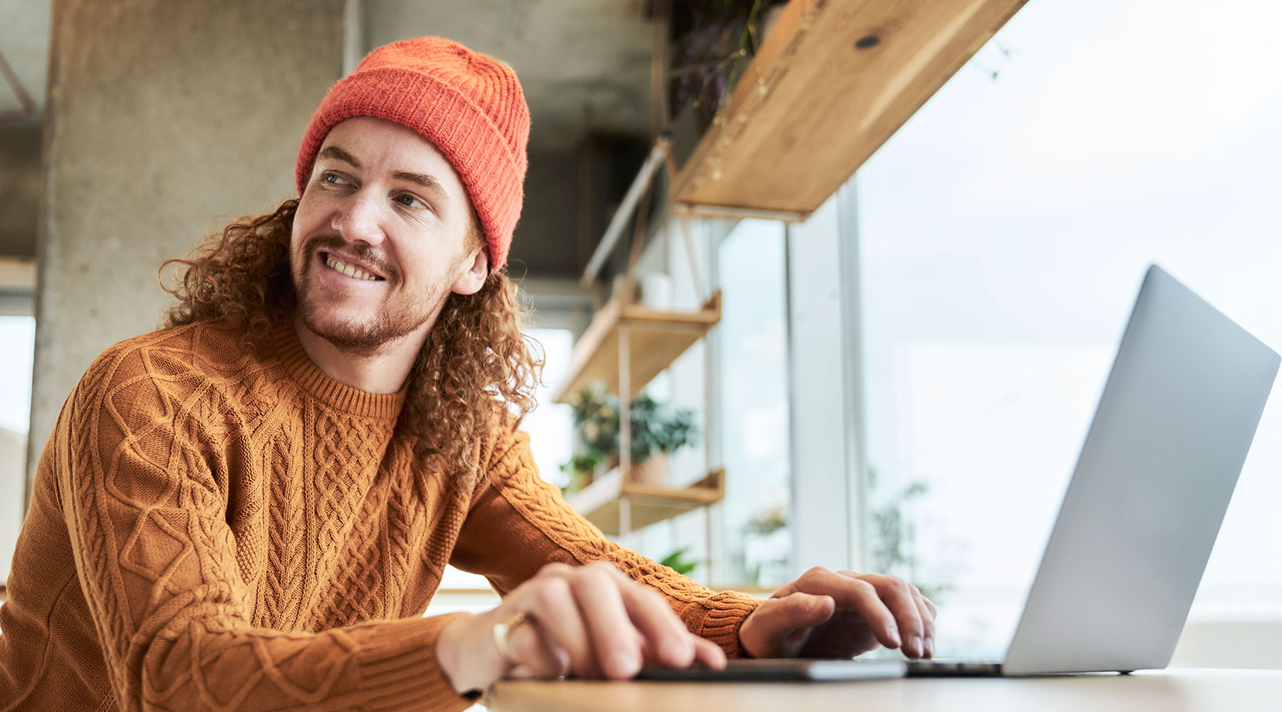 Eine Person mit einem orangefarbenen Pullover und einer passenden Mütze arbeitet an einem Laptop. Der Raum ist modern gestaltet mit großen Fenstern und Pflanzen im Hintergrund.