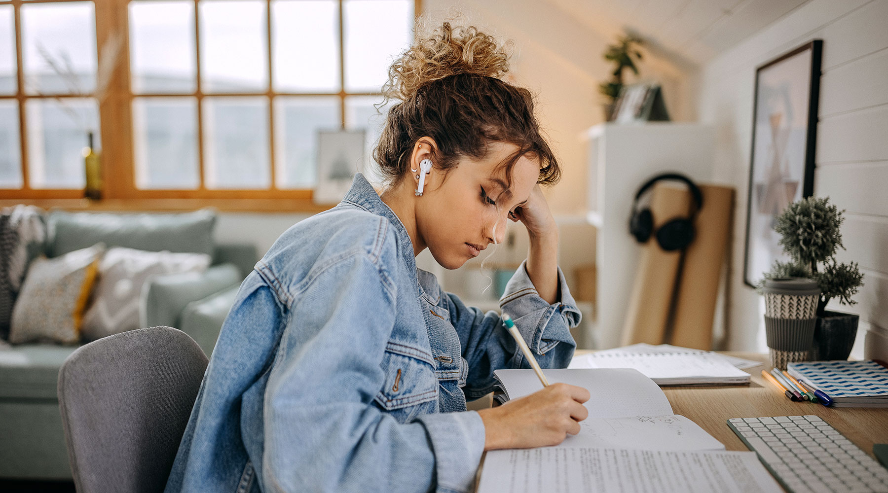 Eine junge Frau sitzt an einem Schreibtisch in einem gemütlichen Raum, schreibt in ein Notizbuch und trägt ein Jeansjacke. Auf dem Tisch befinden sich Pflanzen, Kopfhörer und Schreibutensilien. Im Hintergrund ist ein Sofa und ein großes Fenster zu sehen.