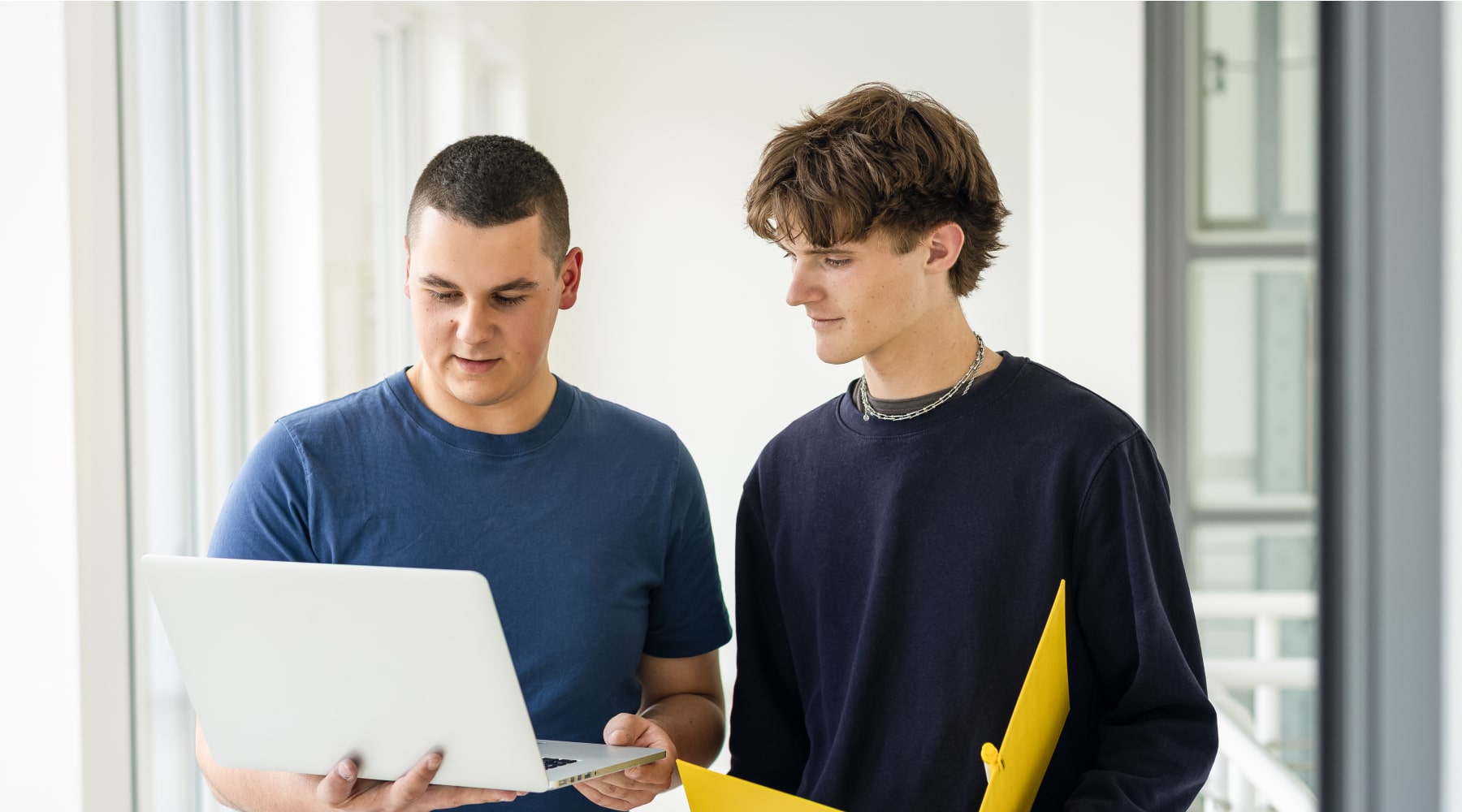 Zwei Personen stehen in einem hellen Raum und betrachten gemeinsam einen Laptop, einer hält eine gelbe Mappe