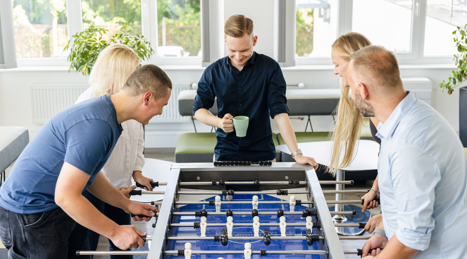 Fünf Personen spielen in einem hellen Raum mit Pflanzen an einem Kickertisch, eine hält eine grüne Tasse. Die Atmosphäre wirkt entspannt und gemeinschaftlich.
