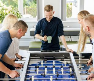 Fünf Personen spielen in einem hellen Raum mit Pflanzen an einem Kickertisch, eine hält eine grüne Tasse. Die Atmosphäre wirkt entspannt und gemeinschaftlich.