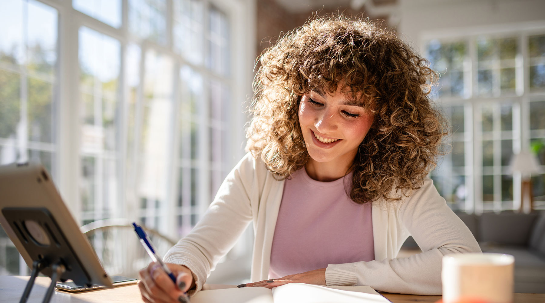 Eine Person schreibt in ein Notizbuch, während sie ein Tablet benutzt. Der Raum ist hell, mit großen Fenstern und natürlichem Licht.