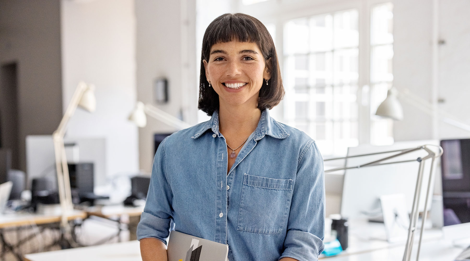 Eine Person in einem Jeanshemd steht in einem modernen Büro und hält ein Tablet. Im Hintergrund sind Schreibtische mit Computern und Lampen zu sehen.