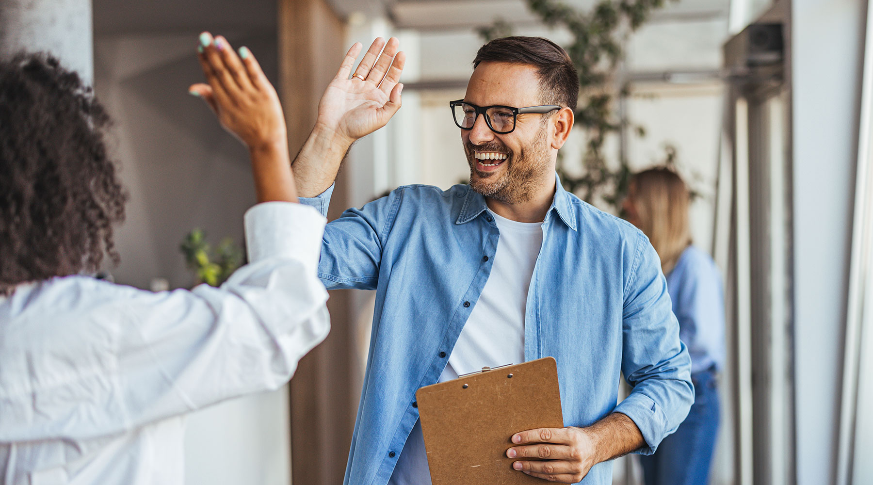 Zwei Personen im Büro geben sich ein High-Five, während eine von ihnen ein Klemmbrett hält. Im Hintergrund sind weitere Mitarbeiter und Pflanzen zu sehen.