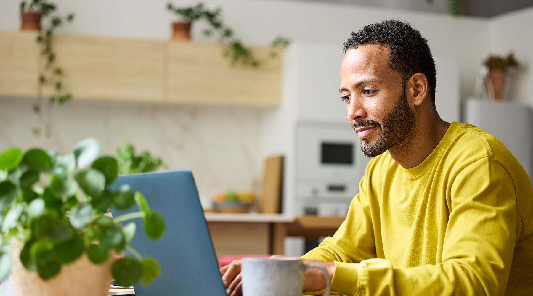 Eine Person in einem gelben Pullover arbeitet an einem Laptop in einer Küche, die mit Pflanzen dekoriert ist. Im Vordergrund steht eine Tasse, und im Hintergrund sind Küchenschränke und ein Ofen zu sehen.