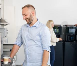 Das Bild zeigt Personen, die Kaffeeautomaten in einem Raum benutzen. Eine Person steht im Vordergrund .
