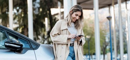 Frau lehnt an ihrem Auto und schaut lachend aufs Handy.