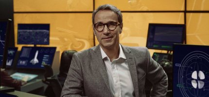 Herr Möller in grauem Anzug steht in einem modernen Kontrollraum. Um sie herum zeigen mehrere Monitore technische Grafiken und Raumfahrtanzeigen, was an eine Missionskontrolle erinnert.