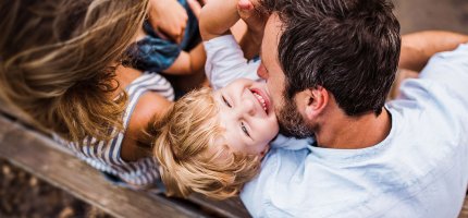 Familie sitzt auf einer Holzbank. Der Vater umarmt den Sohn, der nach oben in die Kamera sieht.