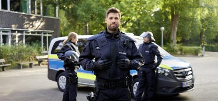Ein lächelnder Polizist mit den Händen an der Schutzweste. Im Hintergrund stehen zwei Kollegen vor einem Polizeiwagen.