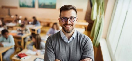Junger Lehrer mit Brille steht vor seiner Klasse und lächelt.