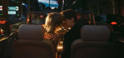 Ein Paar sitzt in einem Cabriolet auf einer belebten Straße bei Dämmerung, umgeben von Stadtlichtern und Scheinwerfern.