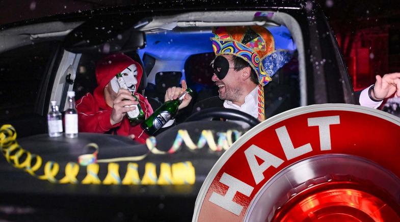 Zwei Personen sitzen in einem Auto, eine trägt eine Maske und hält eine Flasche, die andere einen bunten Hut. Im Vordergrund ist ein "Halt"-Signal zu sehen.