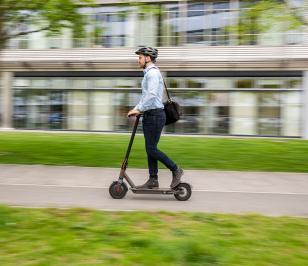 Person fährt auf einem E-Scooter, trägt Helm und Umhängetasche, modernes Gebäude im Hintergrund.