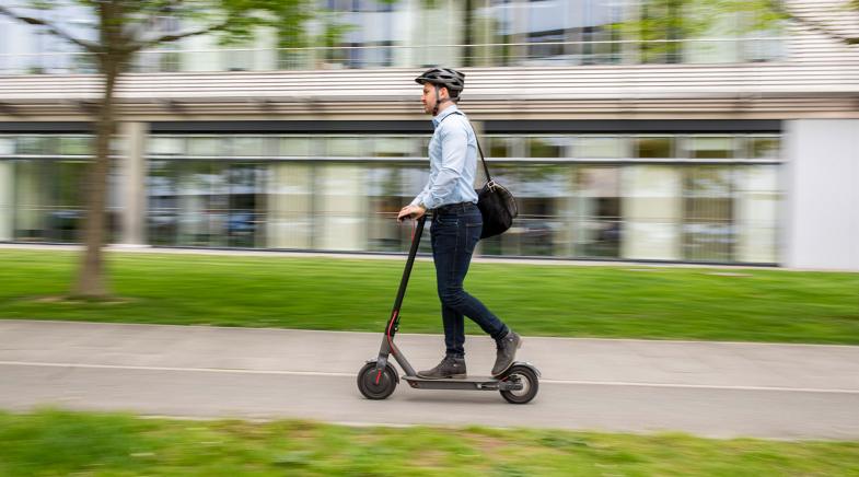 Person fährt auf einem E-Scooter, trägt Helm und Umhängetasche, modernes Gebäude im Hintergrund.