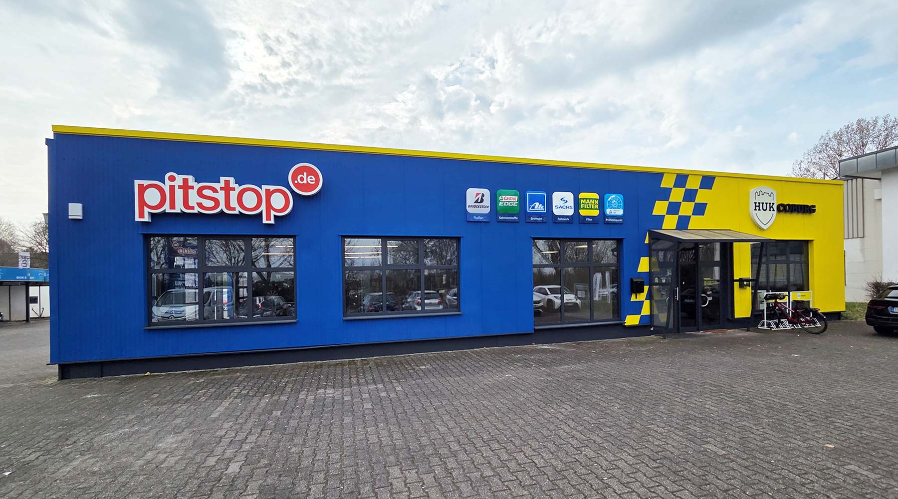 Das Bild zeigt ein blau-gelb gestrichenes Werkstattgebäude mit dem "pitstop.de"-Logo und mehreren Markenlogos.
