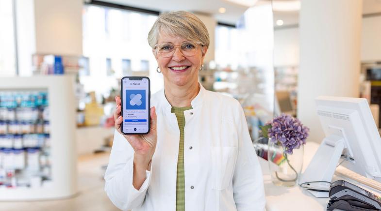 Eine Person in einem weißen Kittel zeigt ein Smartphone mit einer E-Rezept-App in einer Apotheke.