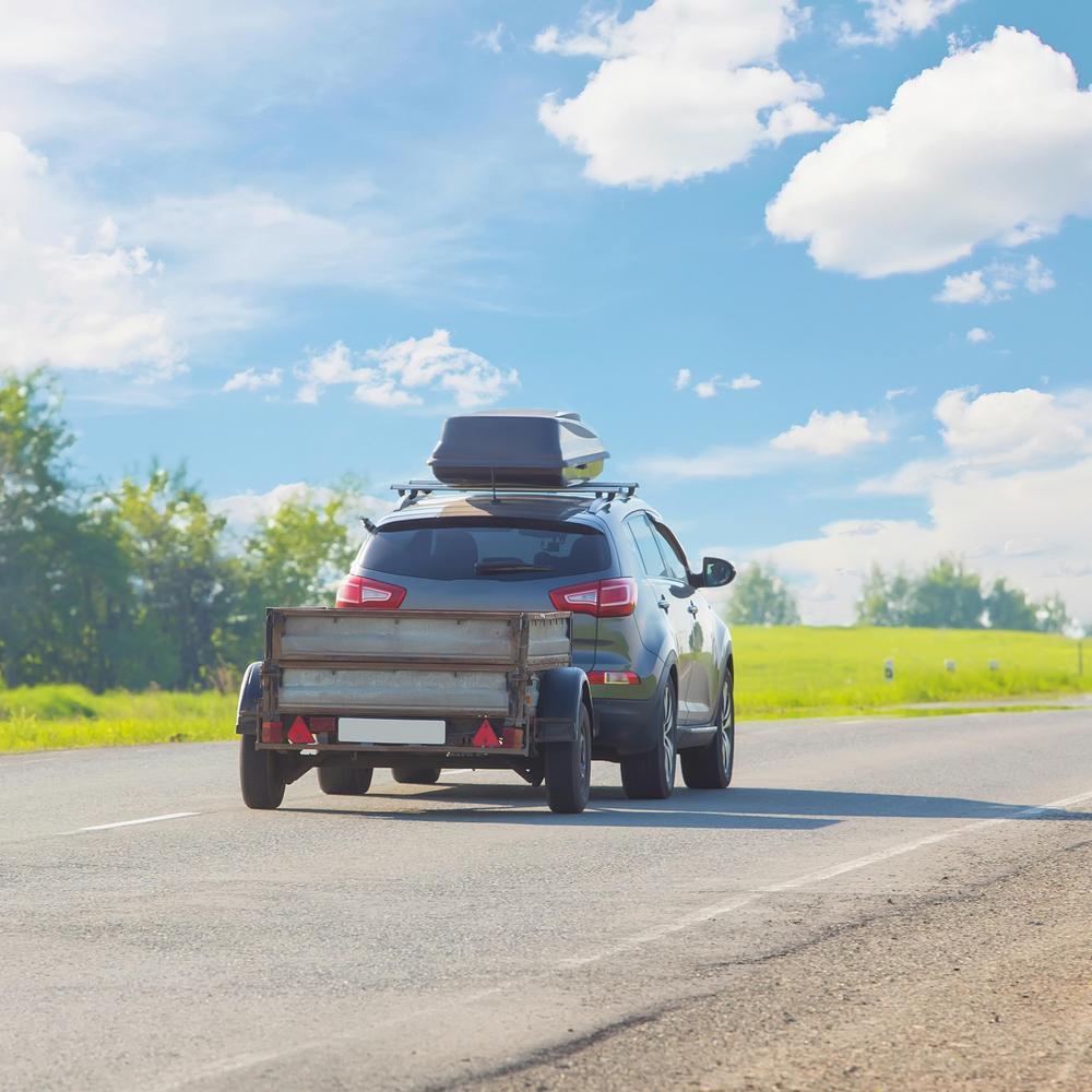 Ein Auto mit Dachbox und Anhänger auf einer Landstraße unter blauem Himmel.