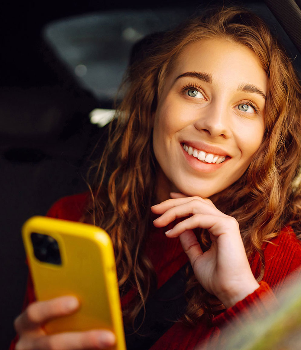 Junge Frau sitzt im Auto und schaut aus dem Fenster, dabei hält sie ein Smartphone mit gelber Hülle in der Hand.