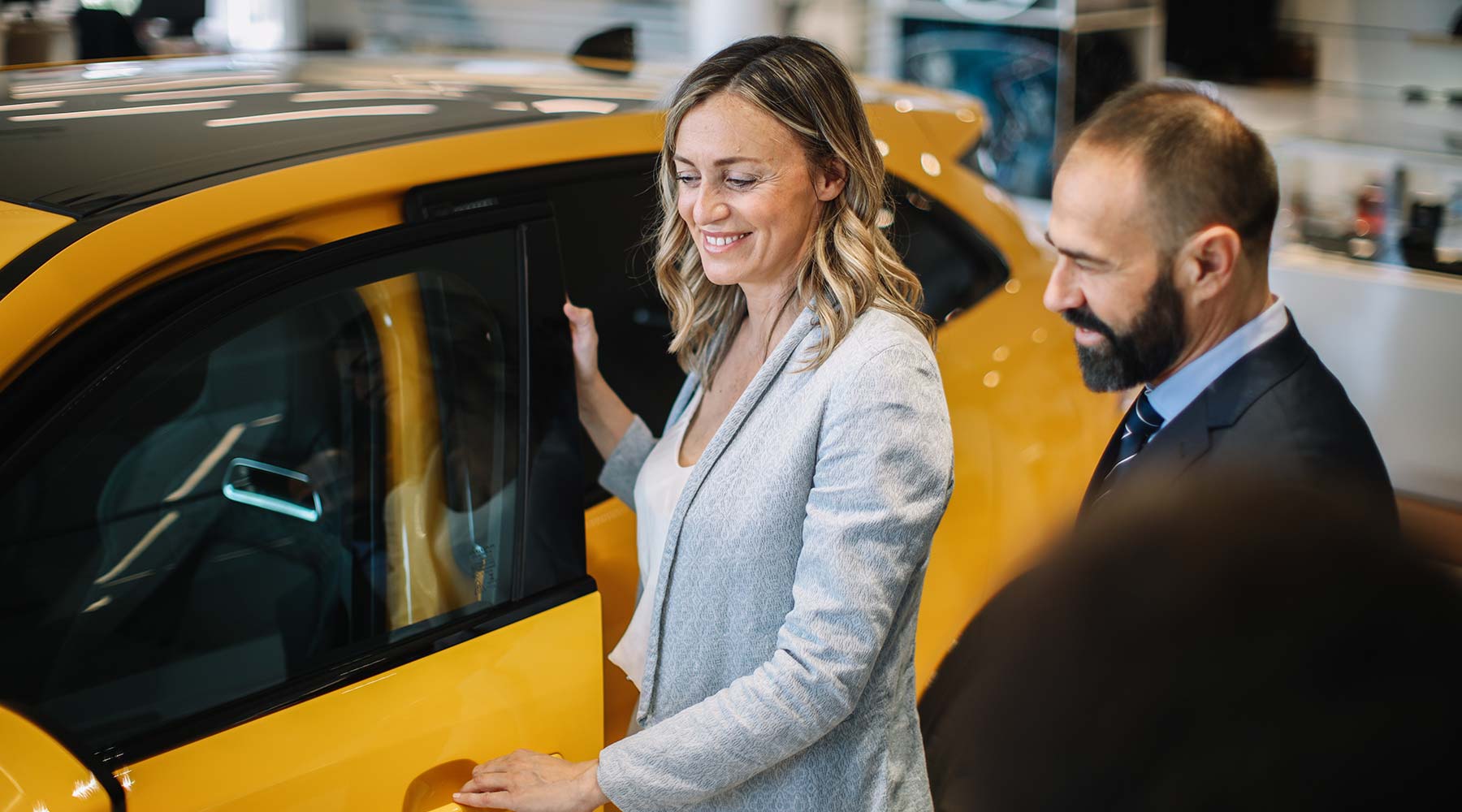 Lächelnde Frau öffnet Tür eines gelben Autos. Daneben steht ein lächelnder Mann.