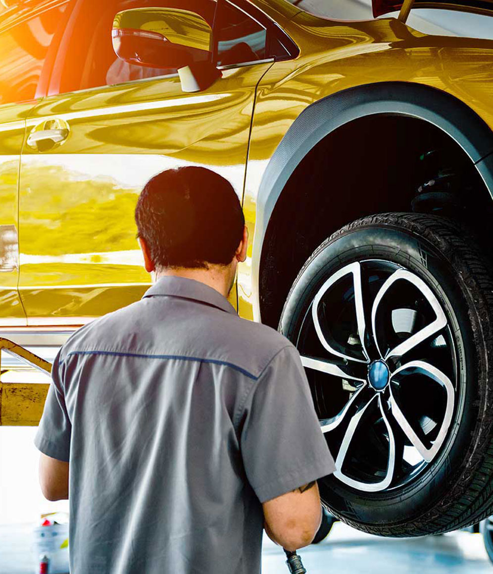 Mann im grauen Shirt schraubt an einem gelben Auto auf einer Hebebühne.